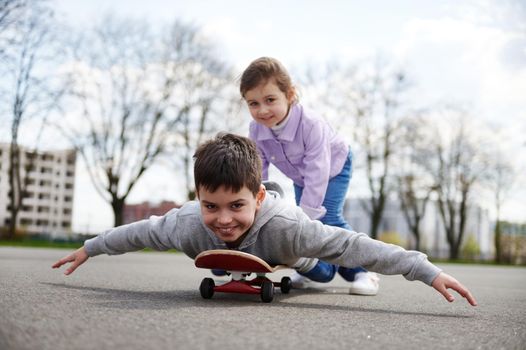 Smiling girl riding her brother on a wooden skateboard enjoying a game together on a sports ground against a background of blurry buildings