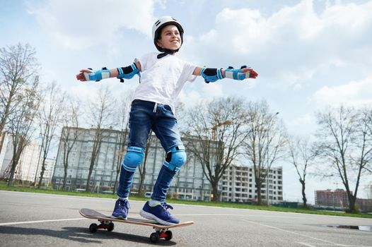 Happy and smiling schoolboy in protective gear and helmet keeps balance while riding a skateboard