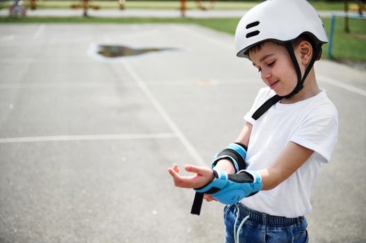 Boy skateboarder wearing safety helmet putting on protective armrests before skating, standing on the playground outdoors