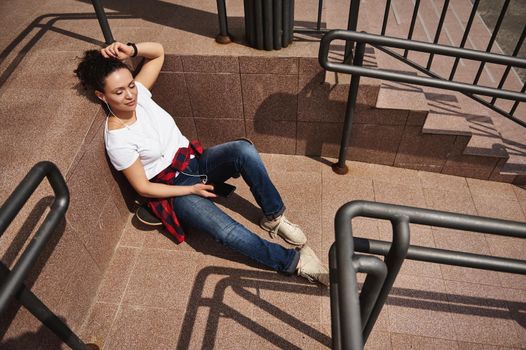 Attractive young woman listening to music on headphones and having rest leaning on steps of unrecognizable building, enjoying warm sunny day