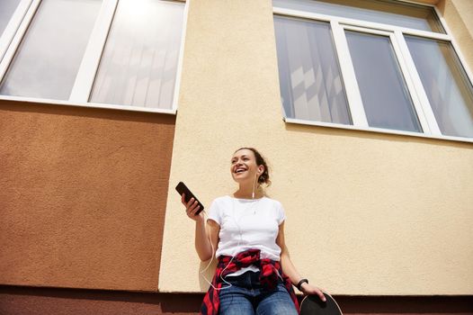 Attractive smiling brunette woman leaning against colored wall posing with skateboard and mobile phone in hands