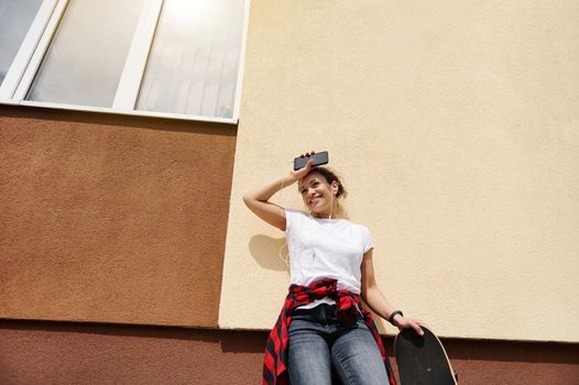 Beautiful hispanic woman in a white t-shirt and jeans posing with a smartphone and a skateboard in her hands against a colored wall on a beautiful sunny day