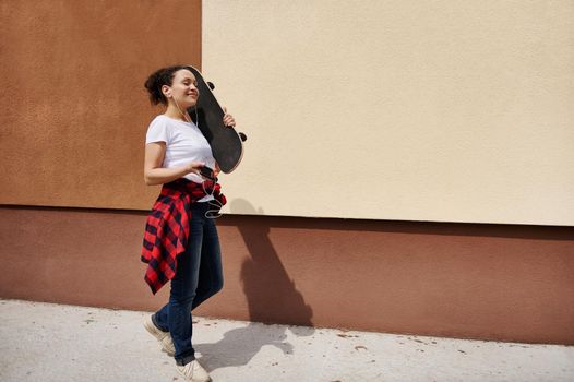 Happy hispanic woman holding wooden skateboard on shoulder walking down the street listening to music against colored wall background