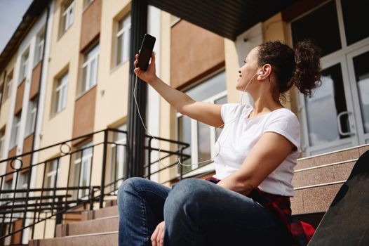 Young African American woman with ponytail and headphones in casual clothing making selfie while sitting on steps