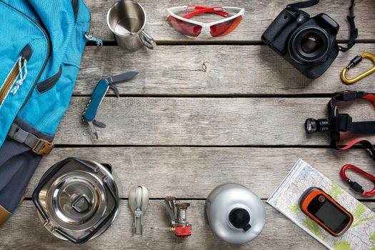 Tourist equipment for travel and tourism on a light wooden floor with an empty space in the middle. Card, knife, rope, carbine, flashlight, shoes, GPS, burner, mug, camera, glasses, backpack.