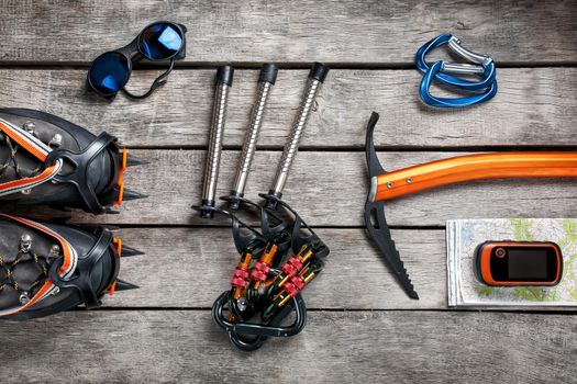 Top view of tourist equipment for a mountain trip on a rustic light wooden floor. Items include glasses, a card, a centipede, a carbine, an ice ax, a GPS, climbing cats.