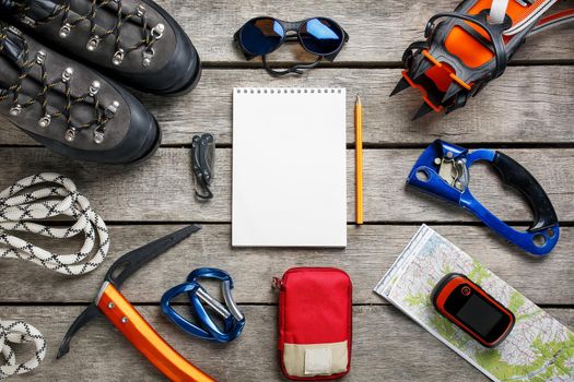 Top view of tourist equipment for a mountain trip on a rustic light wooden floor with a notebook, pencil and empty space in the middle. Items include glasses, a card, a multi-knife, a rope, a carbine, a flashlight, a first aid kit, an ice ax, a GPS, mountaineering cats.