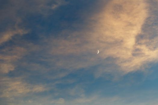 Growing moon in the blue sky among the clouds lit by the sun at sunset. Blue, pink and orange color