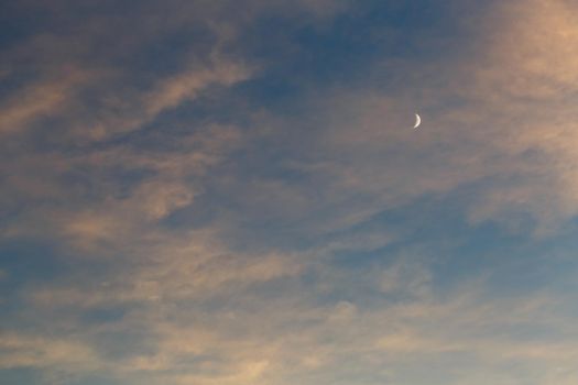 Growing moon in the blue sky among the clouds lit by the sun at sunset. Blue, pink and orange color