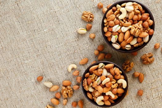 A mixture of cashew nuts, almond nuts, pistachios, hazelnuts and walnuts in a wooden cup against the background of burlap fabric. Nuts as structure and background, macro. Two cups of nuts.