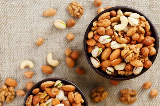 A mixture of cashew nuts, almond nuts, pistachios, hazelnuts and walnuts in a wooden cup against the background of burlap fabric. Nuts as structure and background, macro. Two cups of nuts.