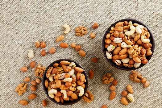 A mixture of cashew nuts, almond nuts, pistachios, hazelnuts and walnuts in a wooden cup against the background of burlap fabric. Nuts as structure and background, macro. Two cups of nuts.