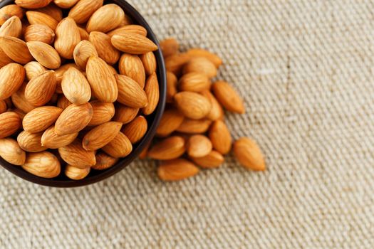 Almonds in a wooden cup on a burlap cloth background. Golden almond closeup in dark brown cup