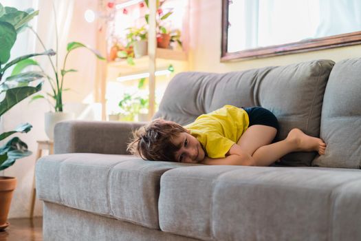 Portrait of happy boy in yellow t shirt lying on sofa, child watching TV and relaxing at home on couch on weekend. Warm and cozy vibes. Home interior with a lot of indoor plants. Smiling Kid fooling.