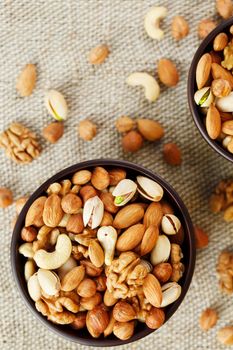 A mixture of cashew nuts, almond nuts, pistachios, hazelnuts and walnuts in a wooden cup against the background of burlap fabric. Nuts as structure and background, macro. Two cups of nuts.