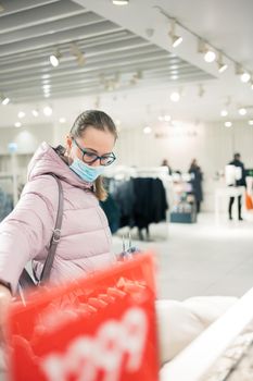 Woman wearing protective face mask shopping clothes in retail shopping store. Concept of lifestyle during corona virus pandemic.