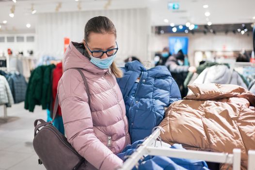 Woman wearing protective face mask shopping clothes in retail shopping store. Concept of lifestyle during corona virus pandemic.