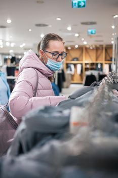 Woman wearing protective face mask shopping clothes in retail shopping store. Concept of lifestyle during corona virus pandemic.