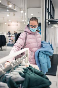 Woman wearing protective face mask shopping clothes in retail shopping store. Concept of lifestyle during corona virus pandemic.
