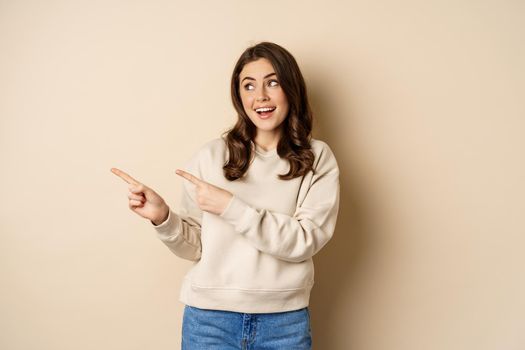 Cheerful attractive female model pointing fingers left, showing store advertisement, banner or logo, standing in sweater over beige background.