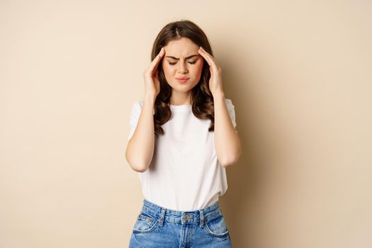 Health and women concept. Portrait of girl feeling sick, touching head temples, feeling headache, migraine, standing over beige background.