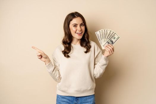 Smiling beautiful woman holding money dollars and pointing finger left, showing logo company banner, standing over beige background.