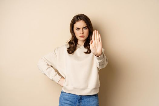 Stop. Serious and confident woman showing extended arm palm, prohibit, forbid smth, blocking something, standing over beige background.