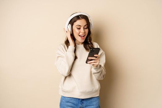 Smiling happy woman listening music in headphones on smartphone, laughing excited, standing over beige background.