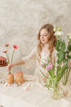 A girl with long hair in a light dress is sitting at the Easter table with cakes, spring flowers and quail eggs. Happy Easter celebration