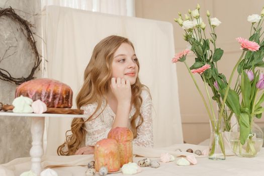 A girl with long hair in a light dress is sitting at the Easter table with cakes, spring flowers and quail eggs. Happy Easter celebration