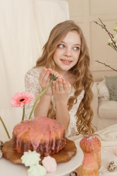 A girl with long hair in a light dress is sitting at the Easter table with cakes, spring flowers and quail eggs. Happy Easter celebration