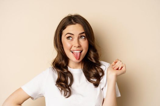 Close up of happy, carefree beautiful woman posing silly, showing tongue, having fun, standing in casual white t-shirt against beige background.