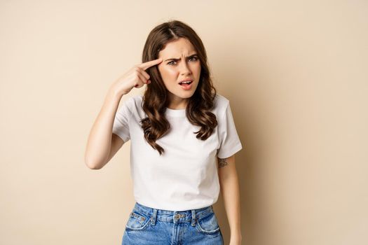Annoyed and angry woman pointing finger at head, scolding someone, standing over beige background.