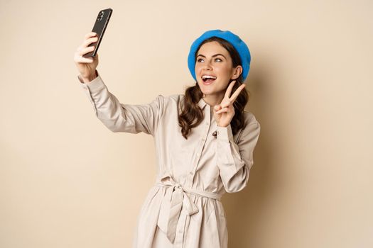 Beautiful modern woman posing for selfie, holding smartphone, taking picture photo on mobile phone, standing happy against beige background.