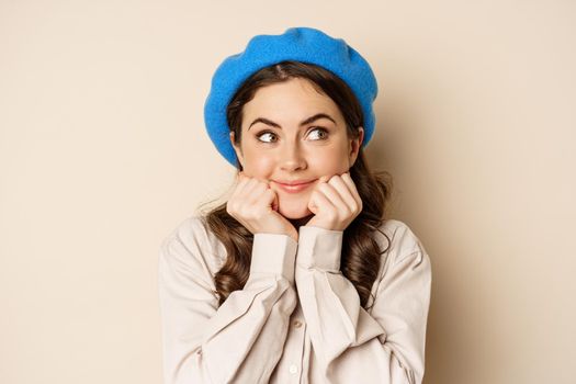 Cute coquettish woman posing, smiling and looking aside with thinking, dreamy face, imaging smth, standing over beige background.