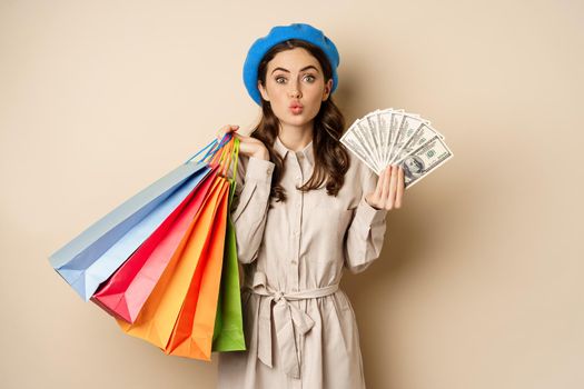 Stylish modern girl feeling satisfaction while shopping, posing with money dollars and shop bags, beige studio background.