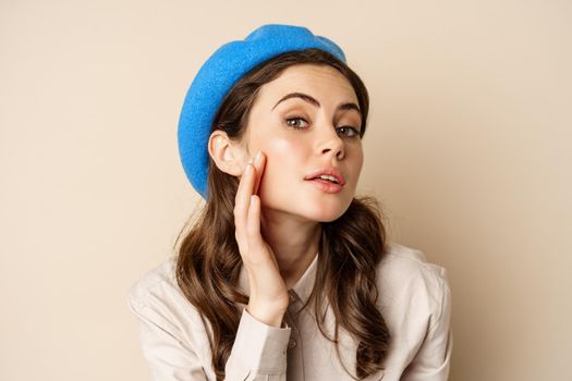 Close up portrait of young beatufiul woman looking in mirror and fixing her makeup, touching soft clear skin, standing over beige background.