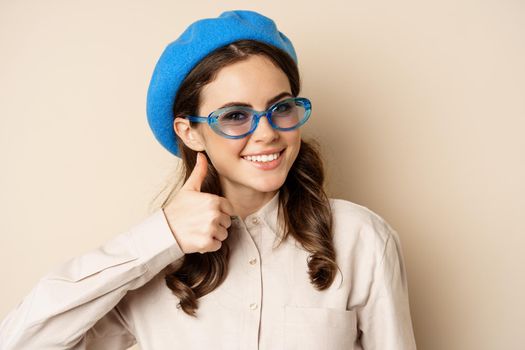Image of happy smiling woman in stylish sunglasses, showing thumbs up in approval, recommending eyewear store, beige background.