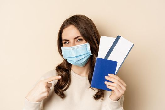 Travel and covid-19 pandemic. Girl in medical face mask travelling, showing passport with two tickets on plane, standing against beige background.