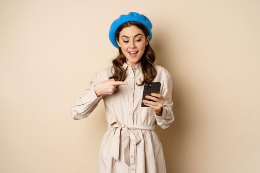 Portrait of stylish modern woman in outerwear, pointing at mobile phone screen and looking happy, smiling pleased, standing over beige background.