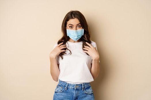 Covid-19 and people concept. Young woman in medical face mask and t-shirt, looking surprised at camera, eyes in disbelief, standing over beige background.