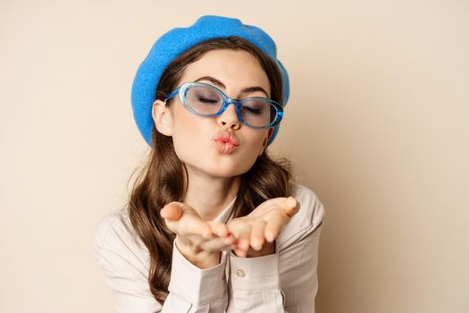 Beautiful stylish woman in sunglasses, blowing air kiss on hands near lips, gazing softly and flirty at camera, posing against beige background.