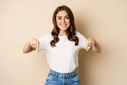 Smiling happy girl pointing fingers down and showing banner, company logo below, click link, standing over beige background.