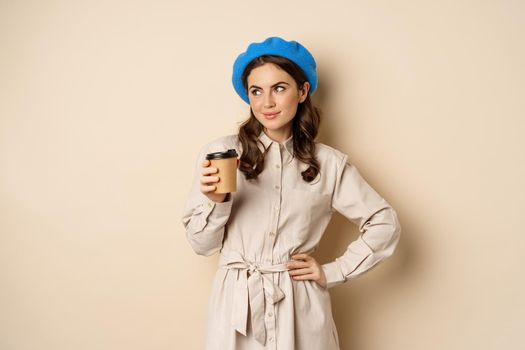 Beautiful happy girl drinking takeaway coffee from cafe and smiling, posing with cup of beverage, beige background.
