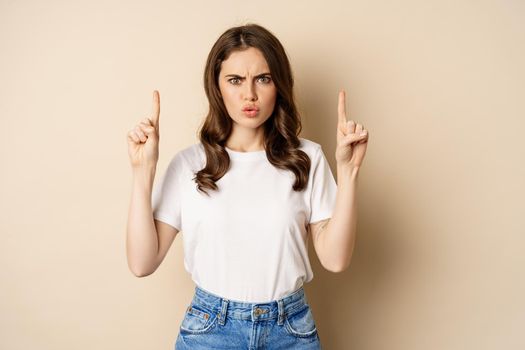 Frustrated young woman pointing fingers up, frowning displeased, standing in white t-shirt over beige background.