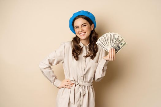 Stylish young woman holding money, cash dollars, smiling and posing satisfied, going on shopping, standing over beige background.