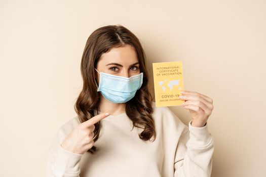 Covid and people concept. Vaccinated woman in face mask showing covid international vaccination certificate, recommend get vaccine, standing over beige background.