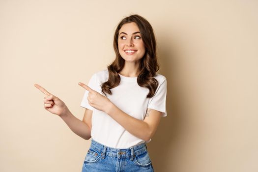 Stylish young caucasian woman smiling, pointing fingers left, showing advertisement, promo offer, standing against beige background.