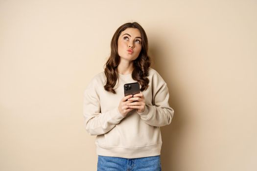 Online shopping. Cute girl thinking while holding mobile phone, imaging smth, daydreaming, standing over beige background.