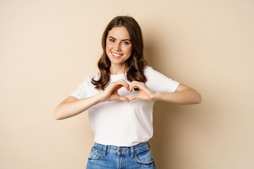 Lovely woman showing heart, love sign and smiling, standing over beige background in white t-shirt, beige background.